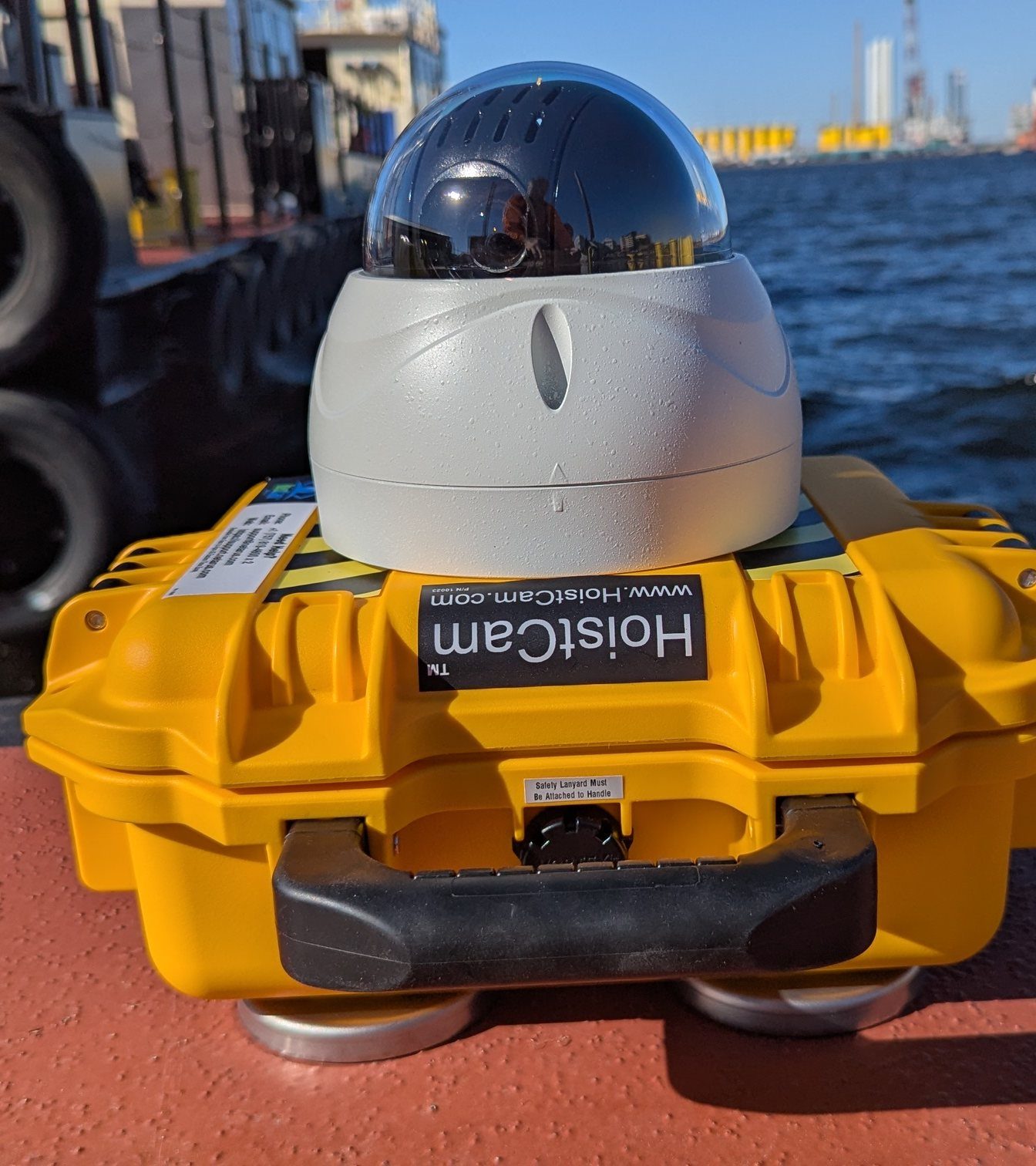 HoistCam HD197 wireless camera in its yellow case on a work barge deck, a tug's fenders and the harbor behind it