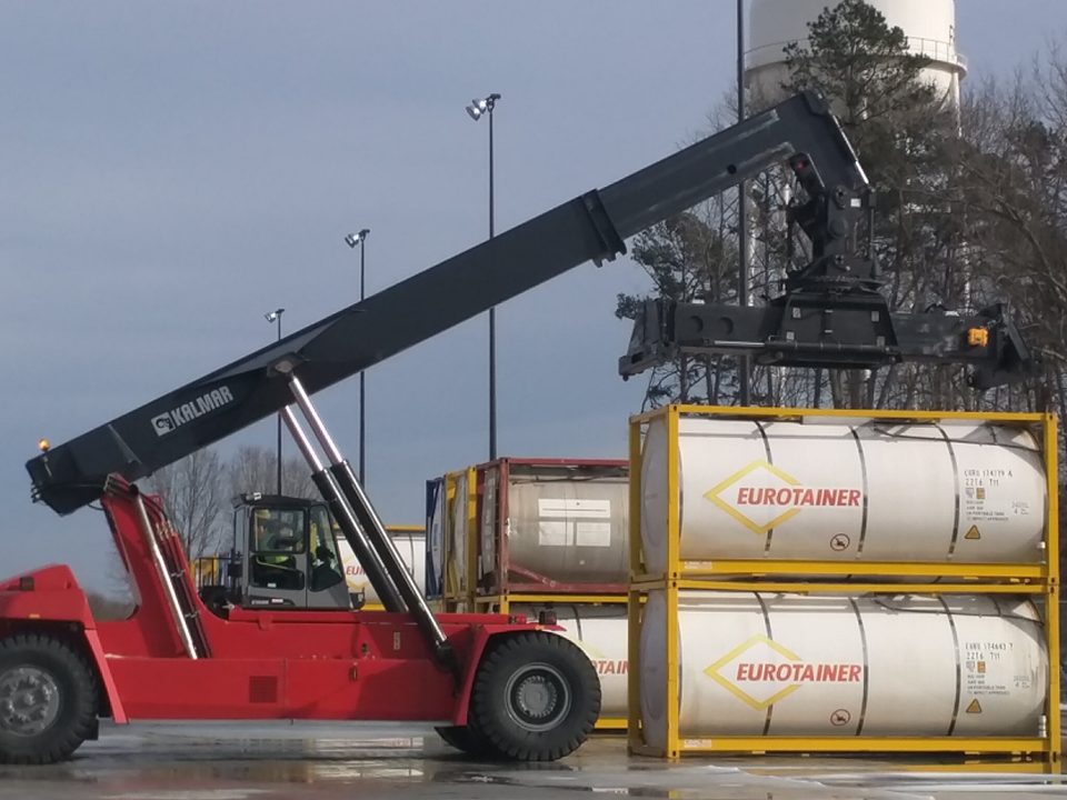 Reach stacker with its boom raised beside stacked tank containers in a yard